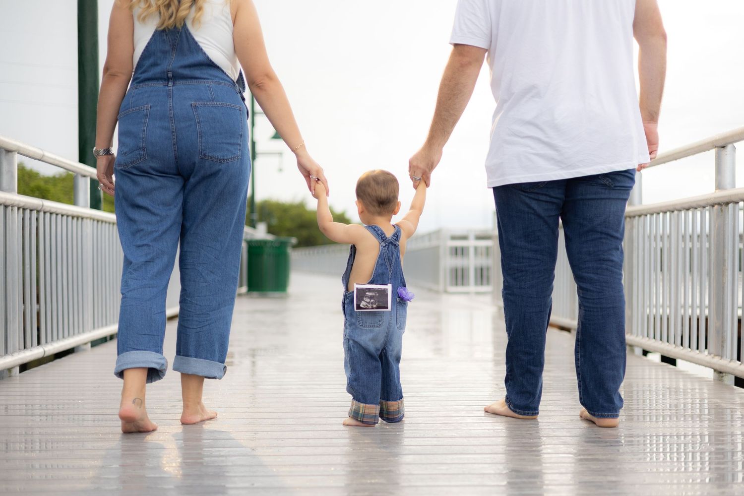 A family of three walks hand-in-hand on a wet boardwalk, with a toddler in denim overalls between two barefoot parents.