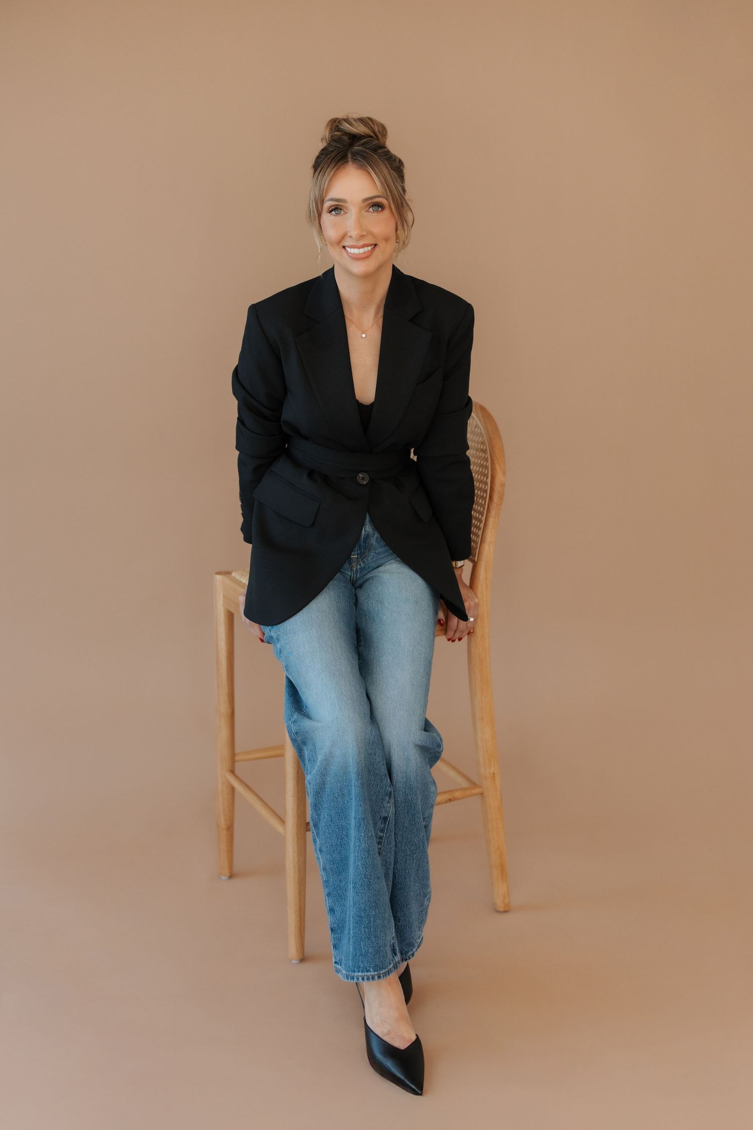 Woman in black blazer and jeans sits on wooden chair, smiling against beige background.
