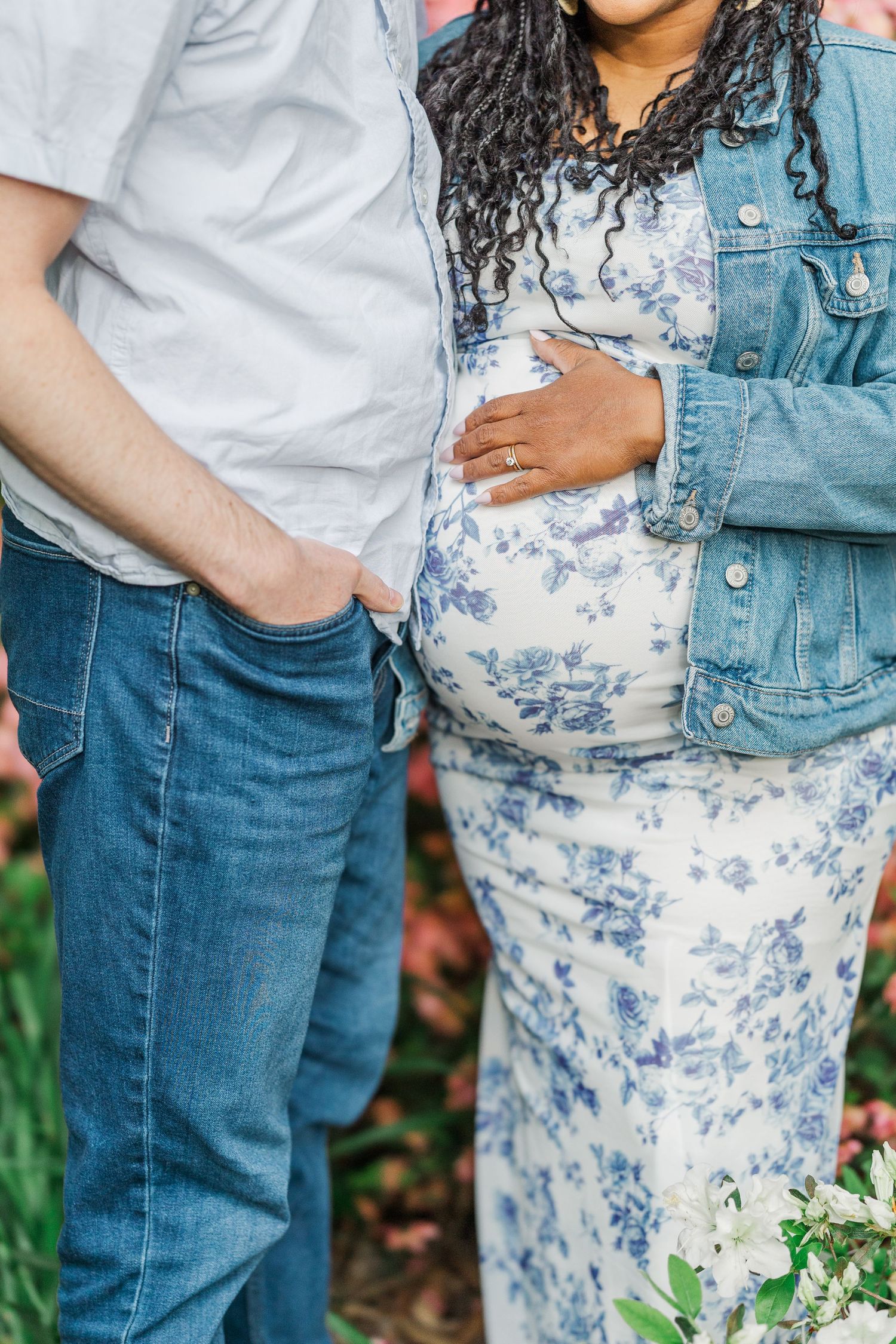 Interracial couple in maternity photo, woman in floral dress holding pregnant belly, man in jeans beside her.