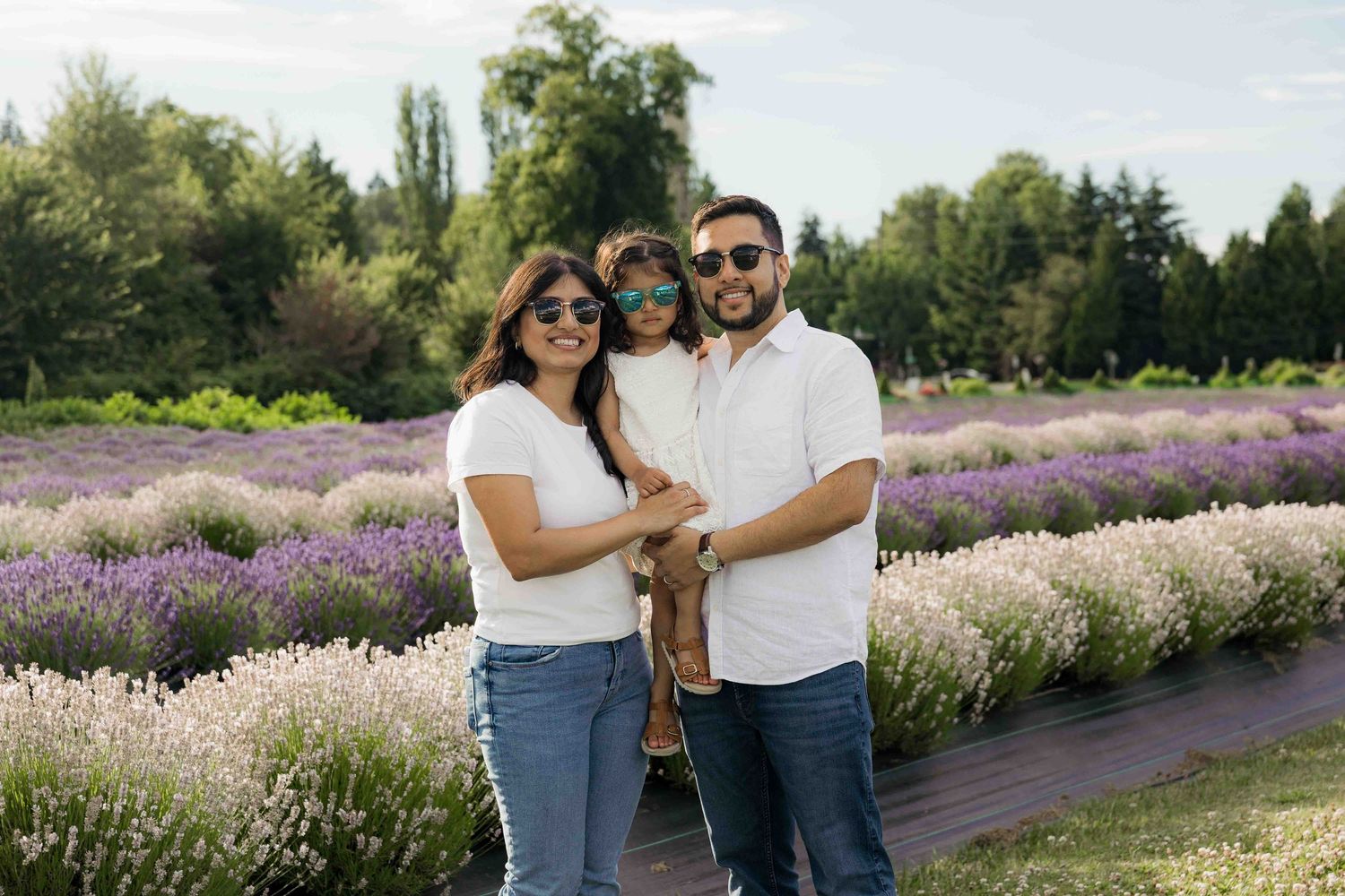 Family of three posing in a lavender field, parents wearing white shirts and sunglasses, holding their young child.