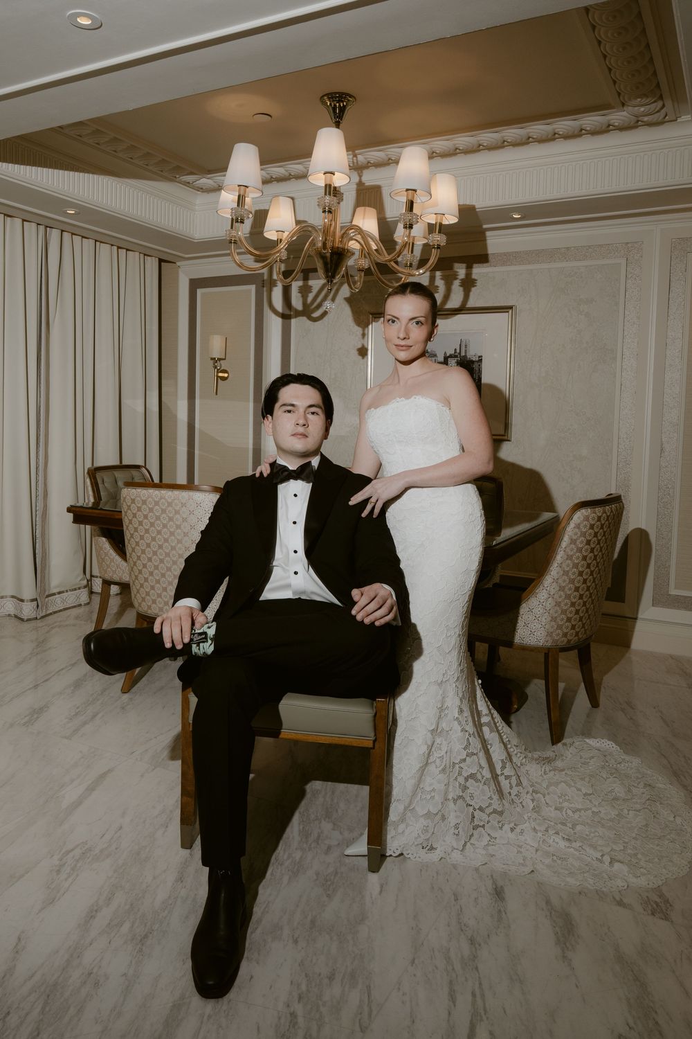Elegant bride in strapless lace gown stands beside seated groom in black tuxedo in a luxurious chandelier-lit room.