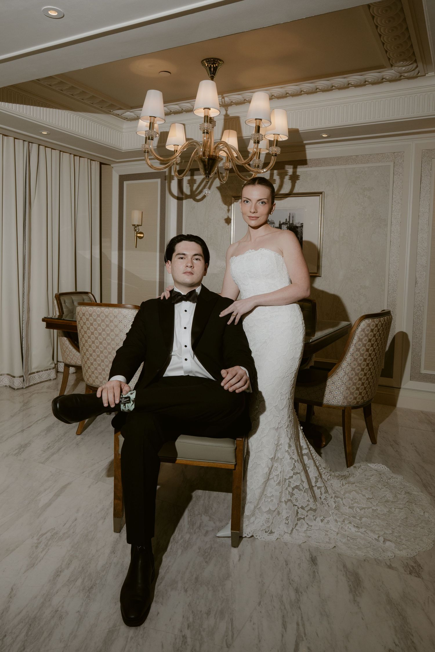 Elegant bride in strapless lace gown stands beside seated groom in black tuxedo in a luxurious chandelier-lit room.