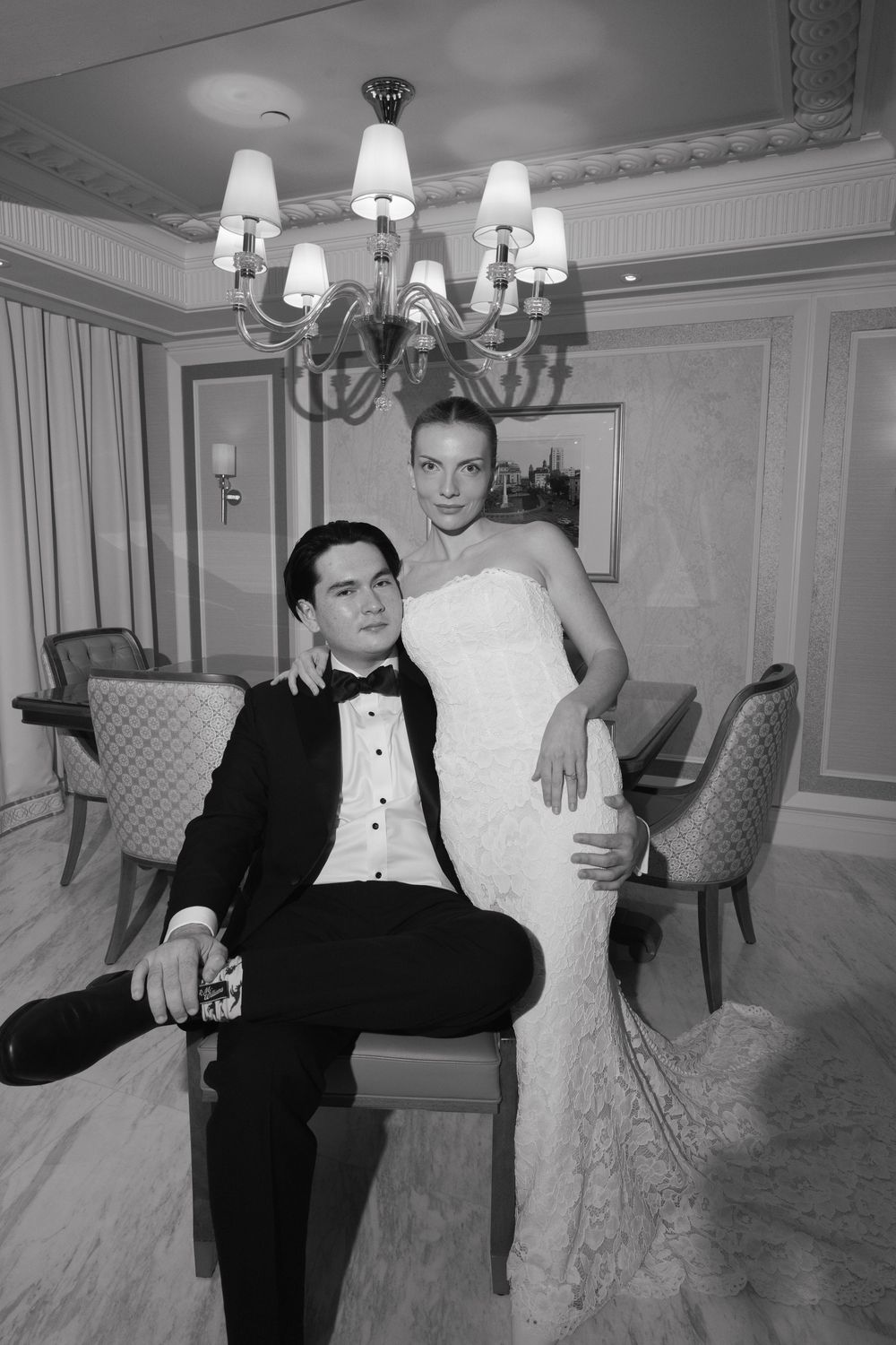 Black and white photo of an elegant bride and groom posing in a luxurious room with chandelier.