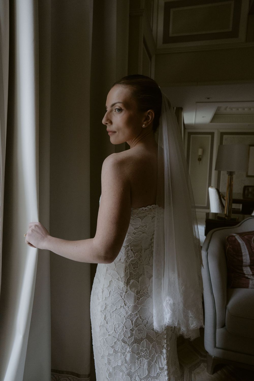 Bride in lace wedding dress and veil gazes out window in elegant room, soft natural light illuminating her profile.