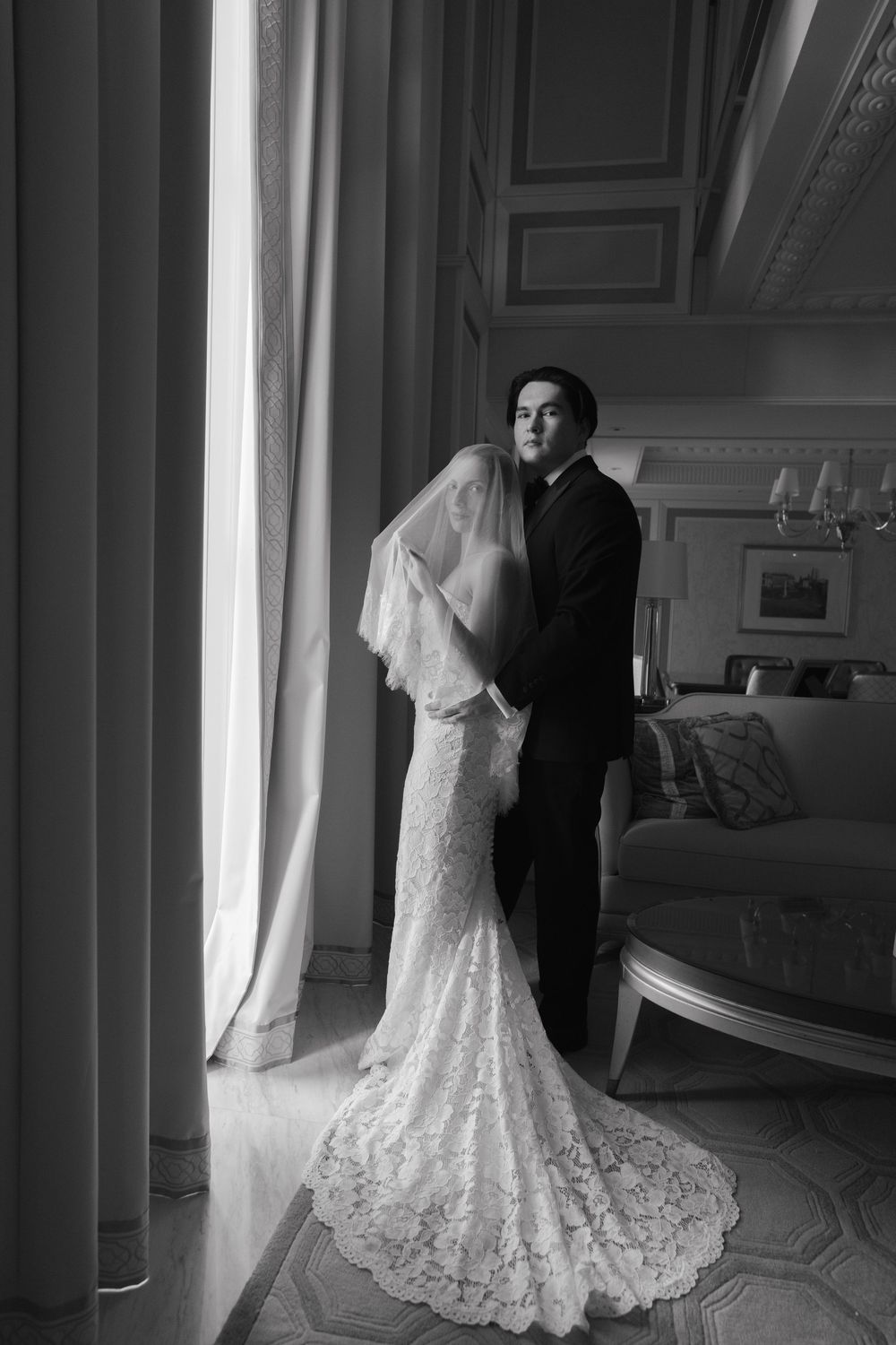Black and white photo of bride in lace gown with veil and groom in tuxedo standing by window in elegant room.