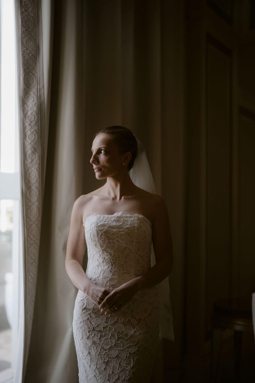 Elegant bride in strapless lace gown and veil gazes out a window in soft natural light.