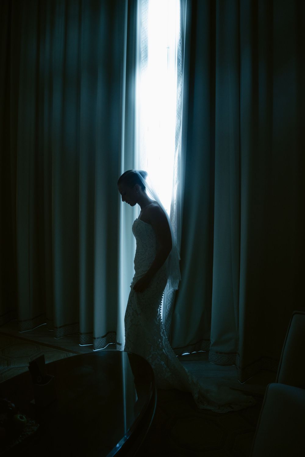 Bride in white gown silhouetted against glowing curtain gap in dark room.