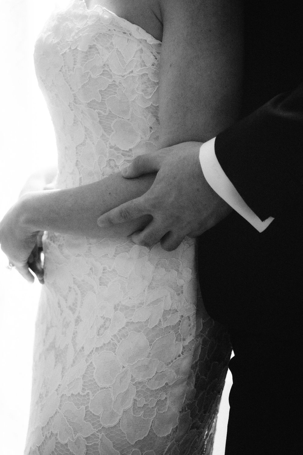 Black and white close-up of bride in lace wedding gown being embraced by groom in suit.