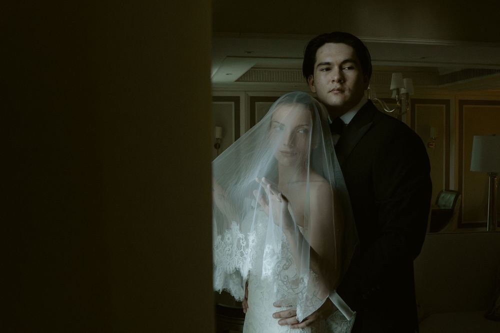 A bride in a white veil and groom in black suit share an intimate moment in a dimly lit room.