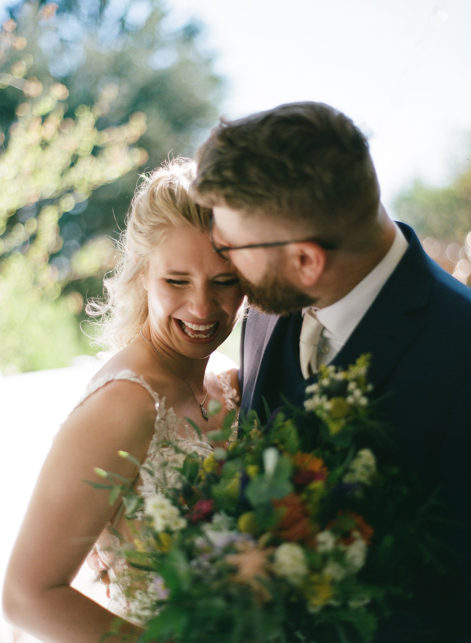 Bride laughing joyfully with groom nuzzling her cheek, holding a lush wildflower bouquet outdoors.