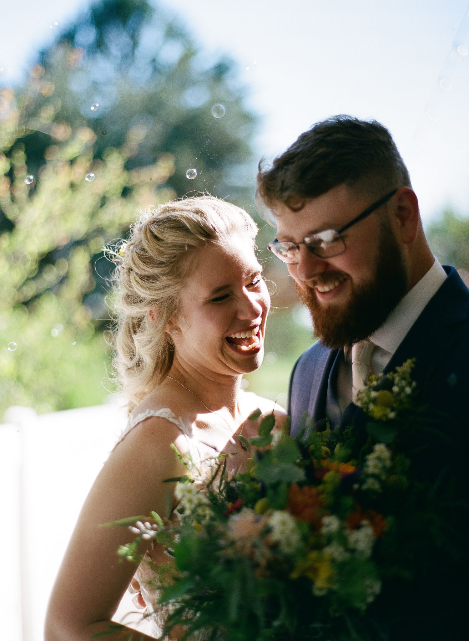Joyful bride and groom laughing outdoors with bubbles, bride holding wildflower bouquet on sunny wedding day.