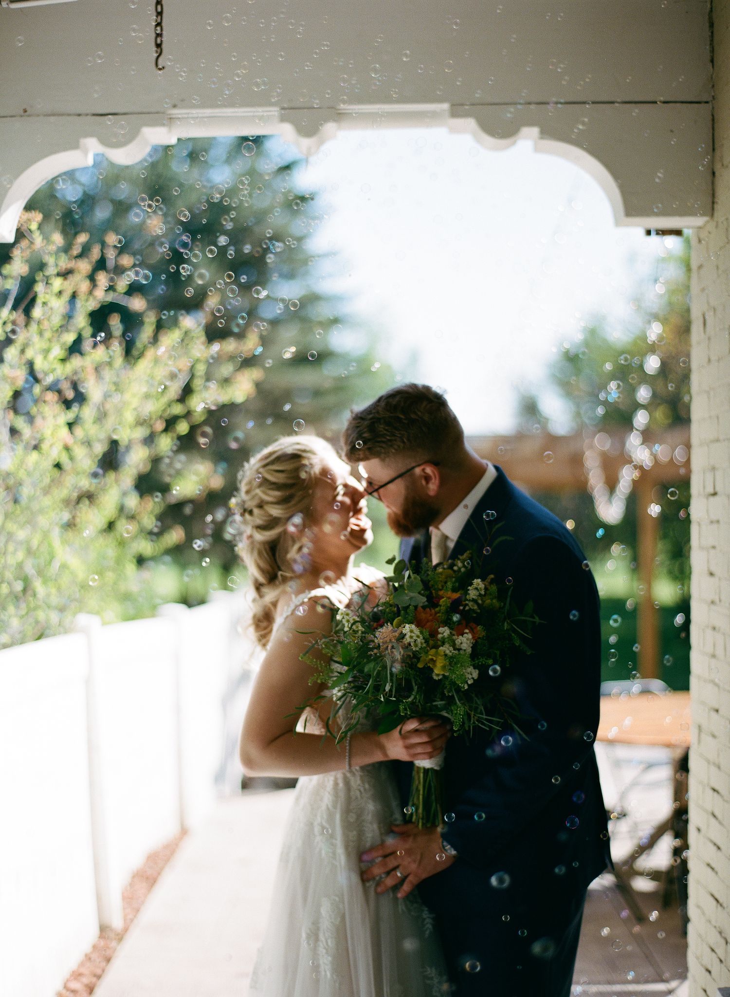Bride and groom share a romantic moment surrounded by bubbles under a white arch with lush greenery.