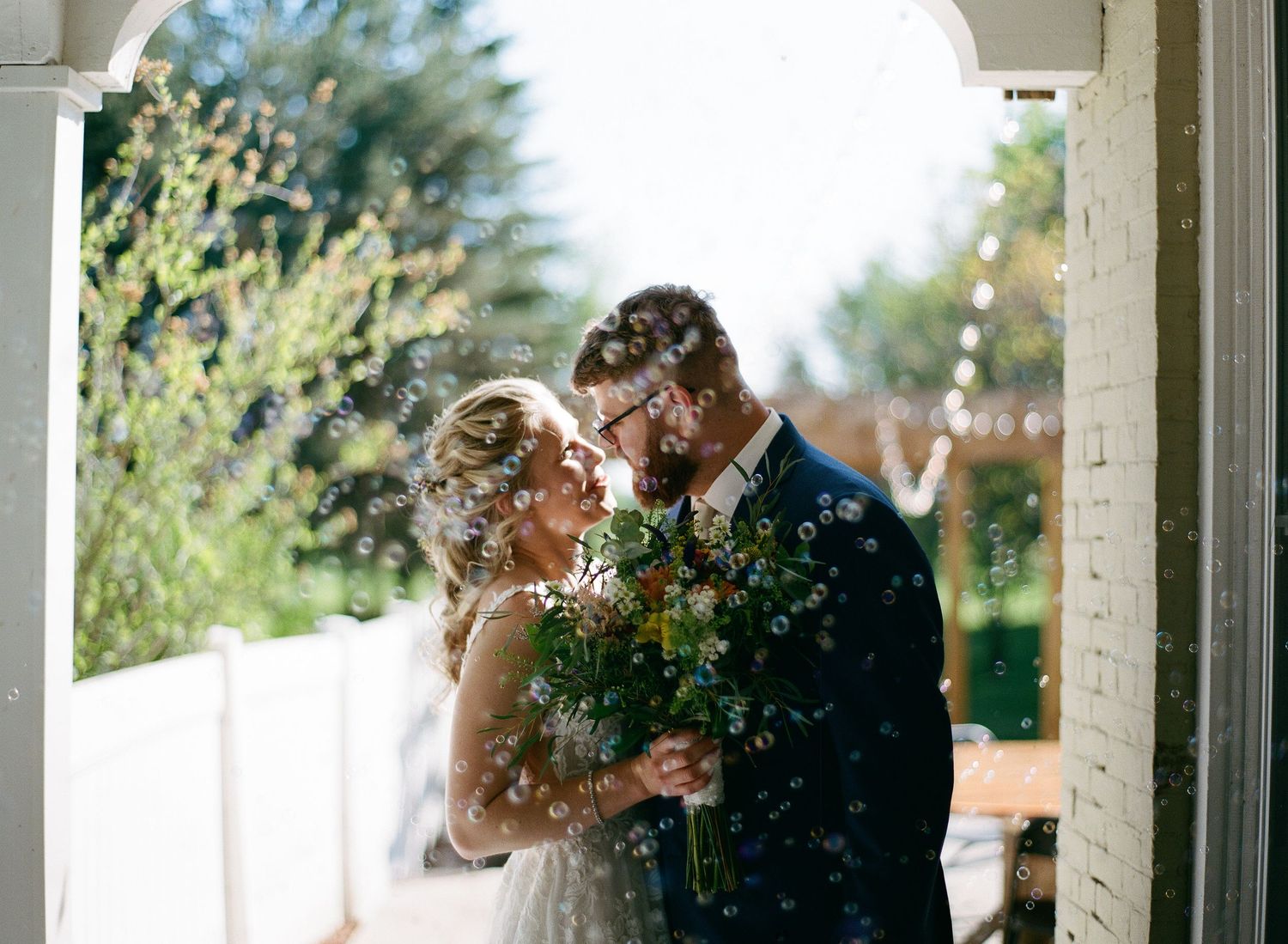 Bride and groom sharing romantic kiss surrounded by bubbles holding greenery bouquet outdoors.