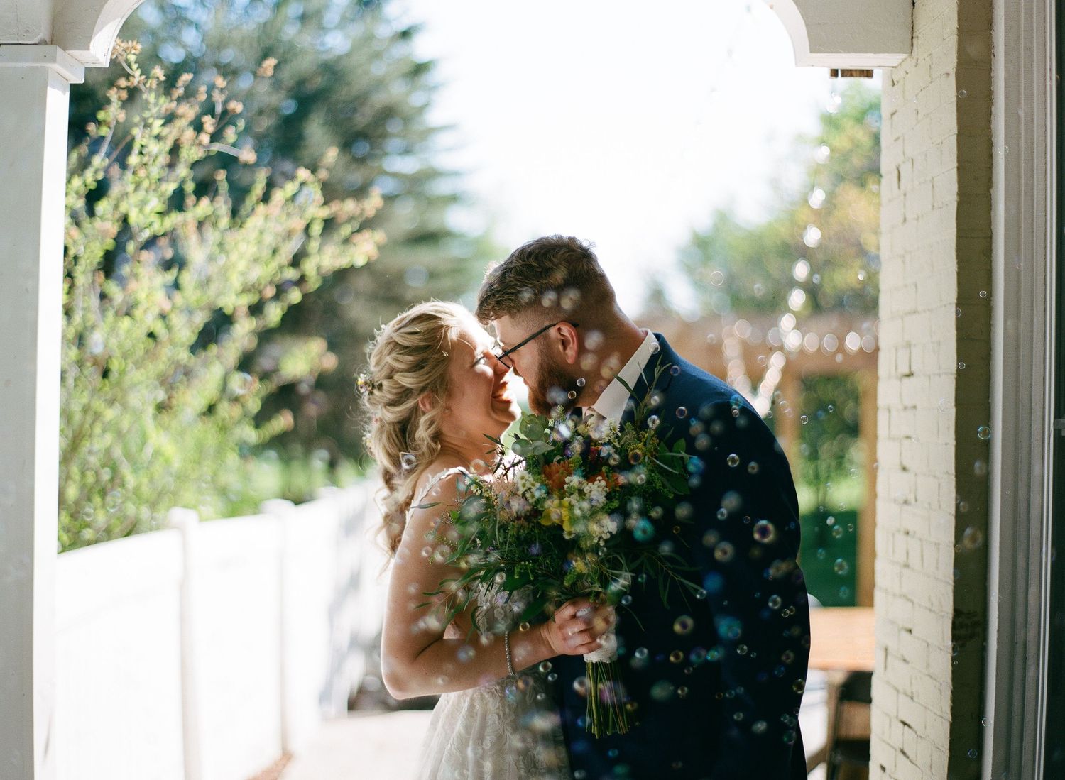 Newlyweds kiss while holding a wildflower bouquet on a sunlit porch with bokeh bubble effects.