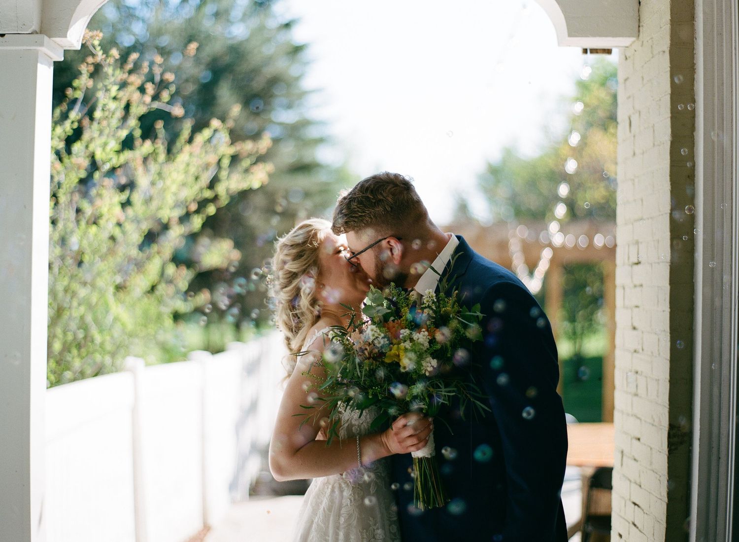 Newlyweds share a romantic first kiss holding a wildflower bouquet in a sunlit doorway with bokeh bubbles.