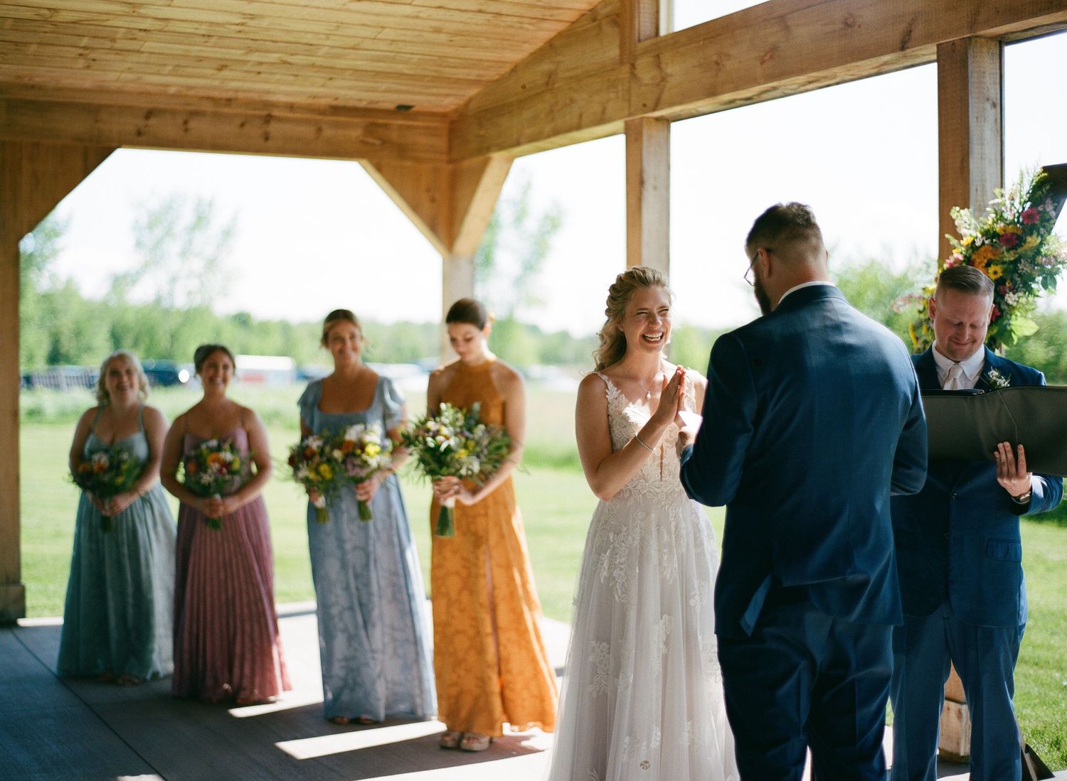 Bride smiles at groom during outdoor wedding ceremony with bridesmaids in colorful dresses holding bouquets under wooden pavilion.