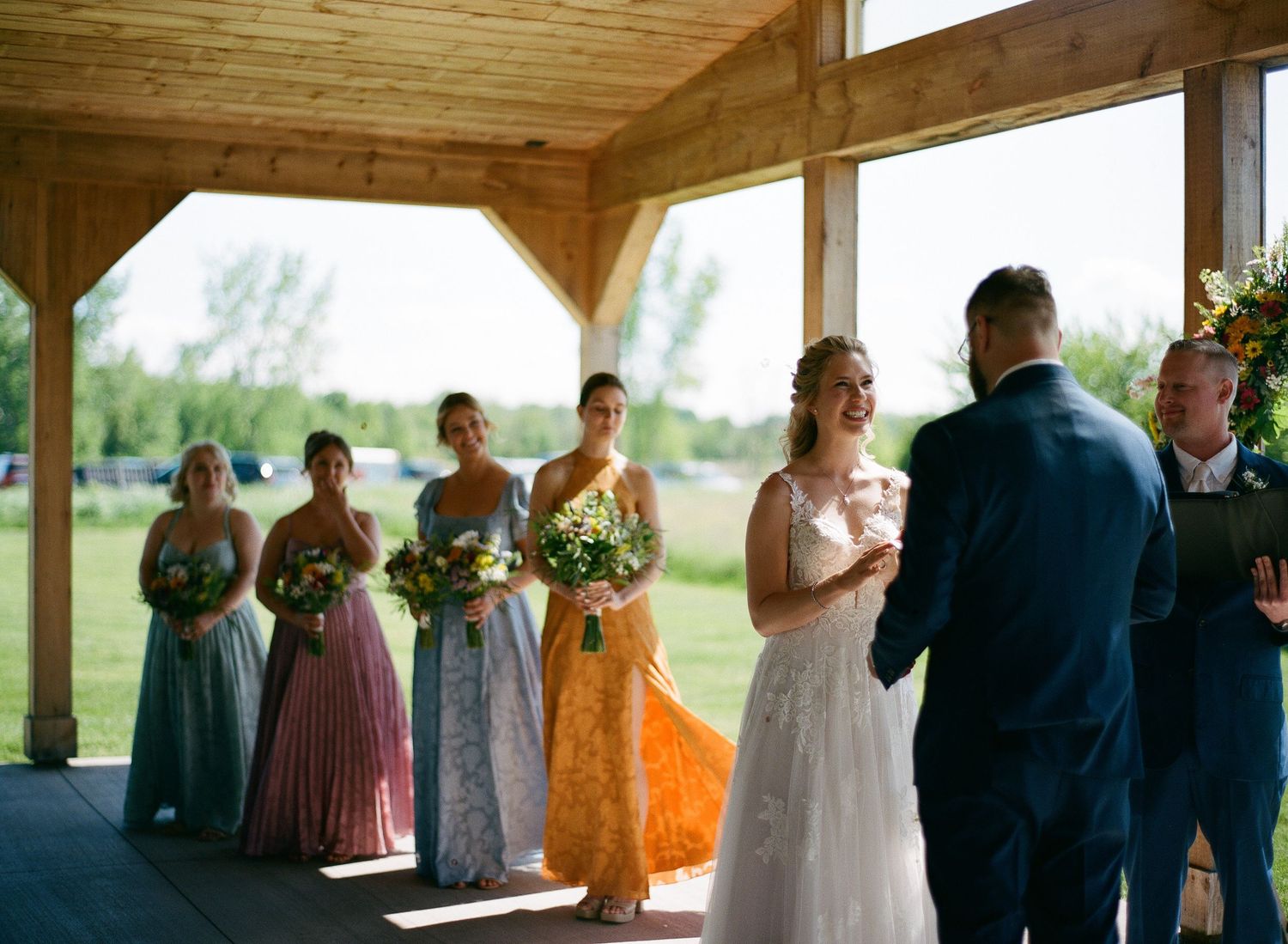 Bride and groom exchange vows under wooden pavilion, surrounded by colorful bridesmaids holding bouquets at outdoor wedding.