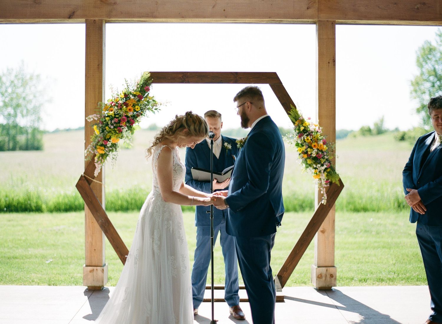 Couple exchanging vows under a hexagonal wooden arch adorned with colorful flowers at an outdoor wedding ceremony.