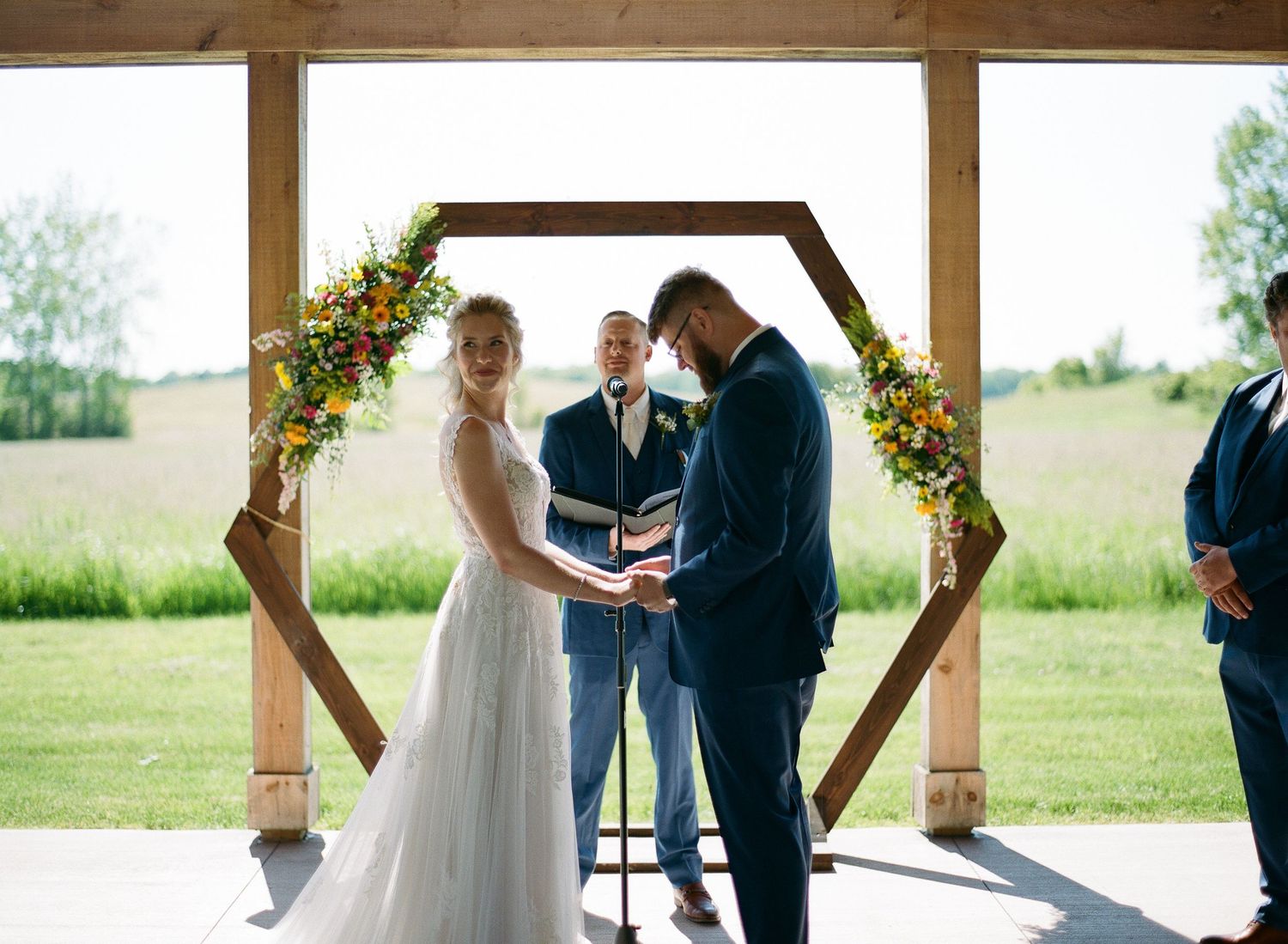 Bride and groom exchange vows under a wooden hexagonal arch adorned with colorful flowers at an outdoor wedding ceremony.
