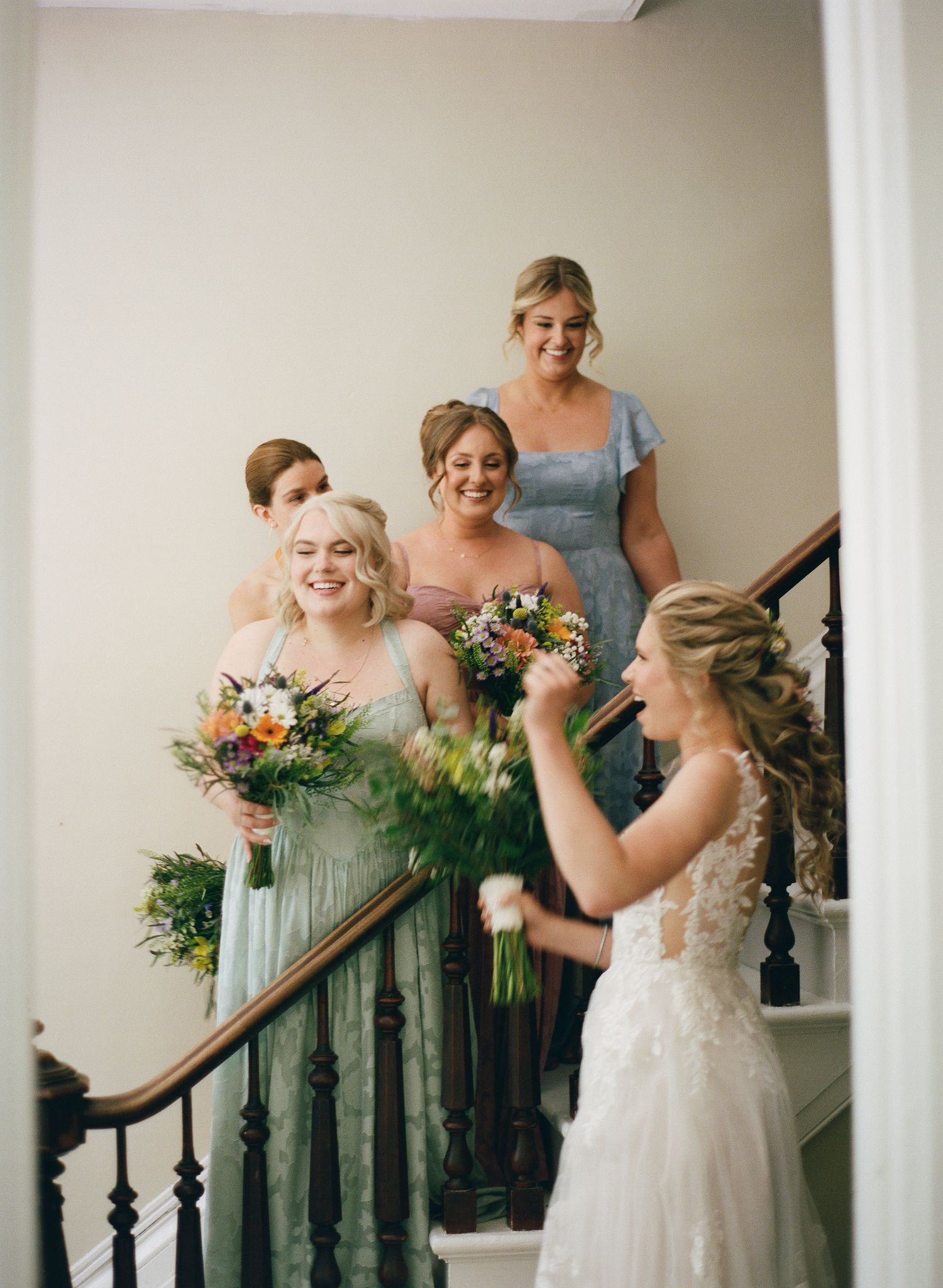 Smiling bridesmaids in sage and dusty blue gowns holding bouquets gather on a staircase with the bride.