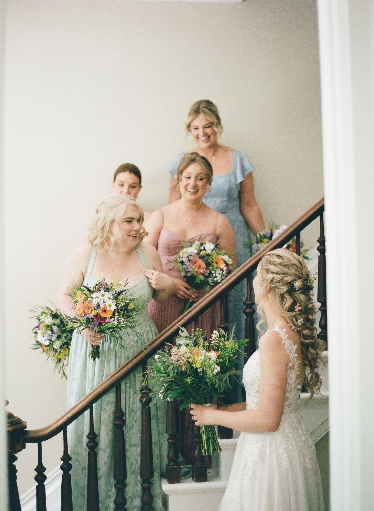 Bride in white lace gown admires bridesmaids in mismatched pastel dresses holding wildflower bouquets on elegant staircase.