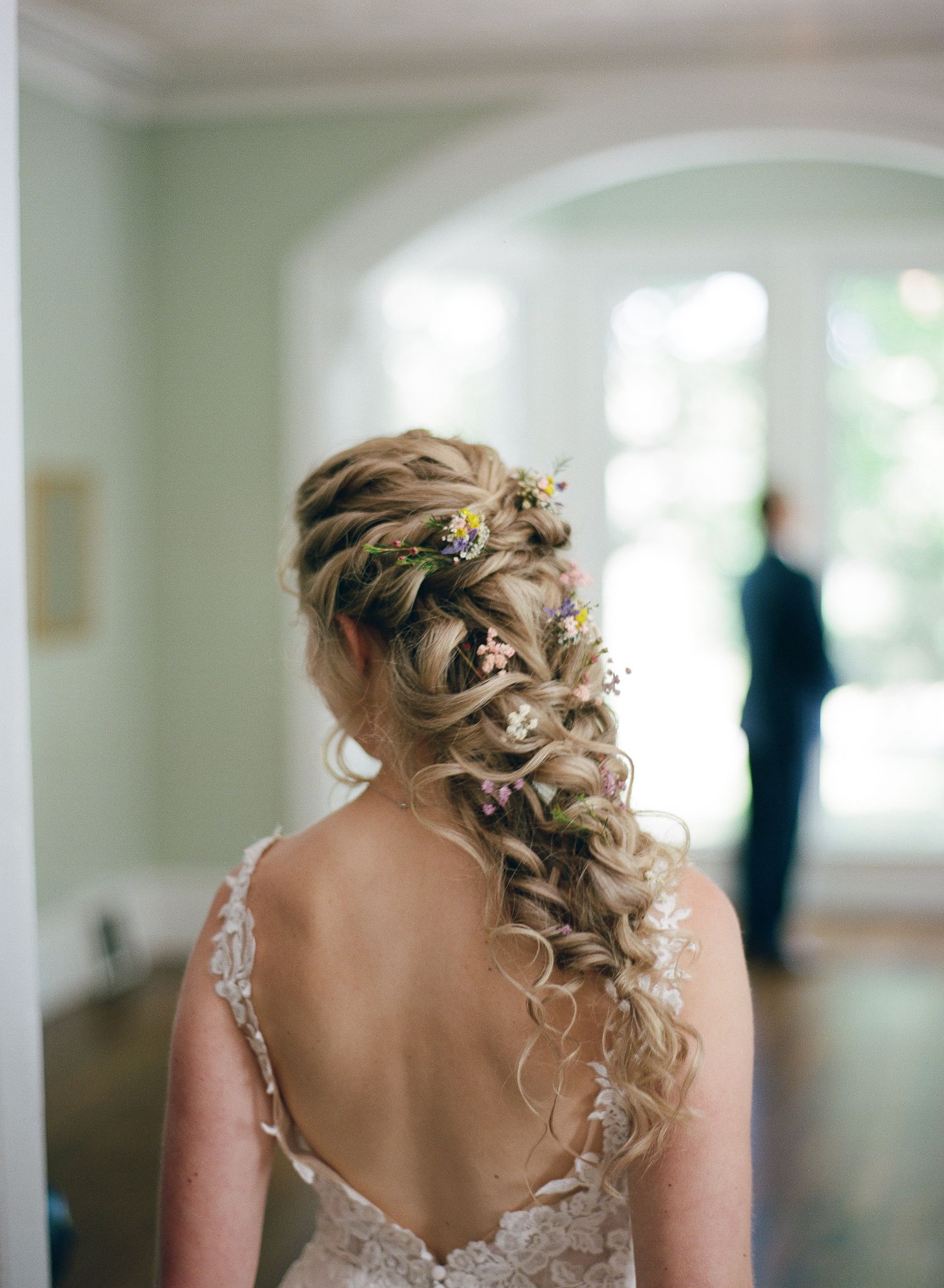 Bride with elegant braided half-up hairstyle adorned with small wildflowers, wearing a lace backless wedding dress.