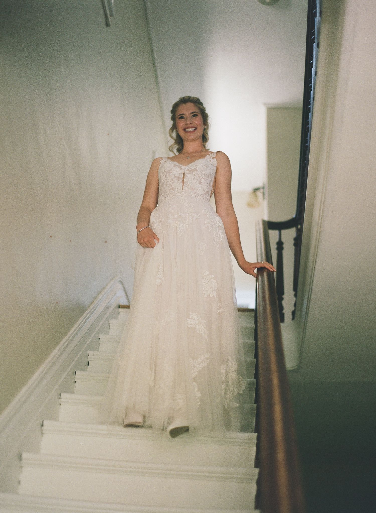 Smiling bride in a flowy lace wedding gown descending an elegant white staircase.