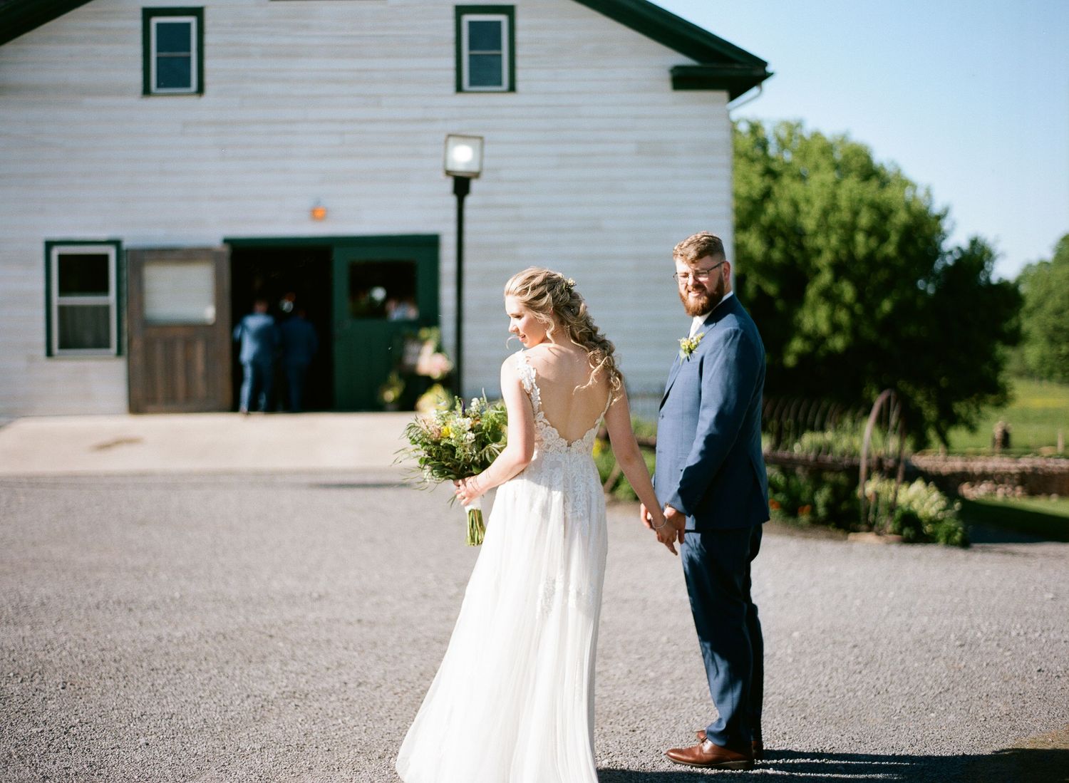 Bride and groom hold hands facing a white barn venue on a sunny summer wedding day.