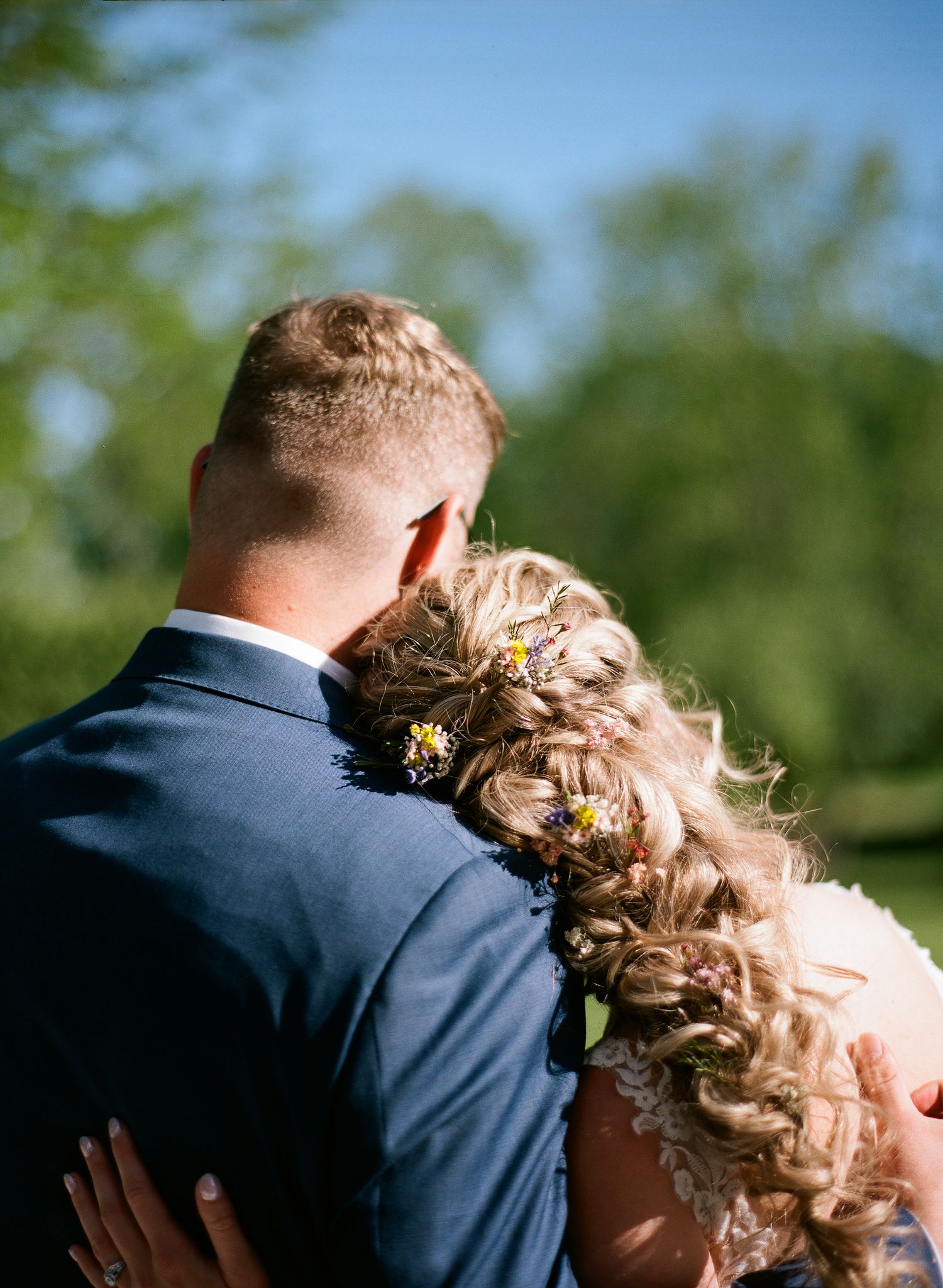 A bride and groom embrace from behind, showing her floral braided updo and his navy suit in sunlight.