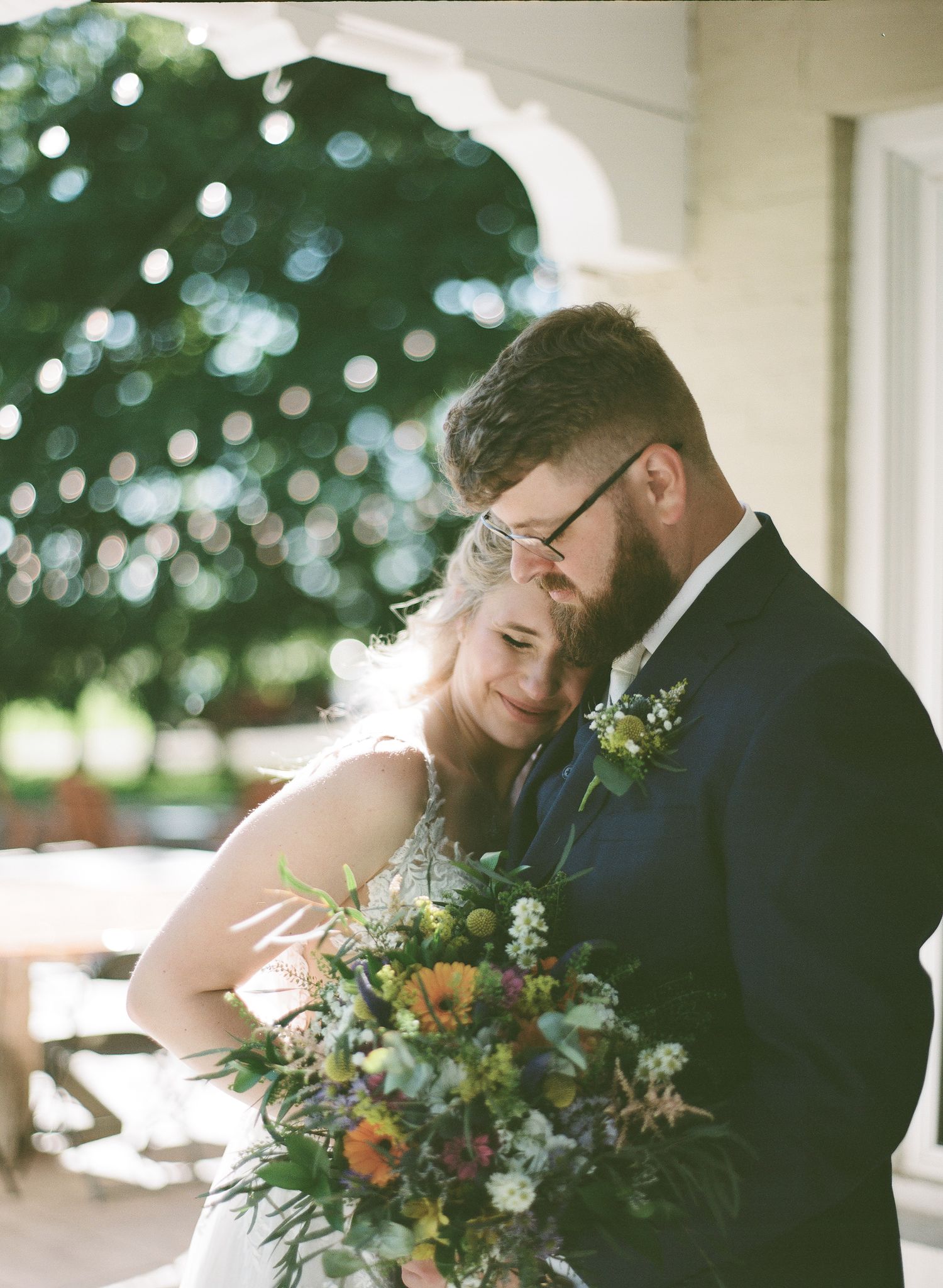 A bride rests her head on the groom's chest while holding a lush wildflower bouquet at an outdoor venue.