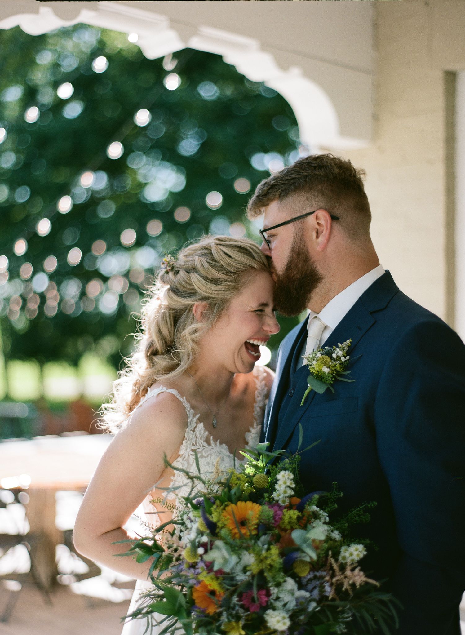 A laughing bride and bearded groom share a joyful moment together holding a vibrant wildflower bouquet.
