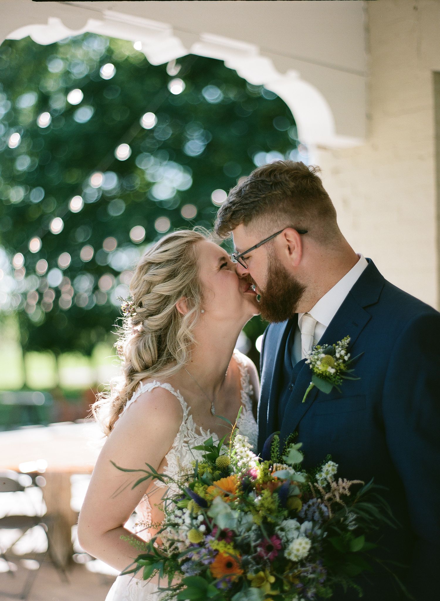 Bride and groom sharing a romantic kiss, bride holding a colorful wildflower bouquet, groom in navy suit with floral boutonniere.