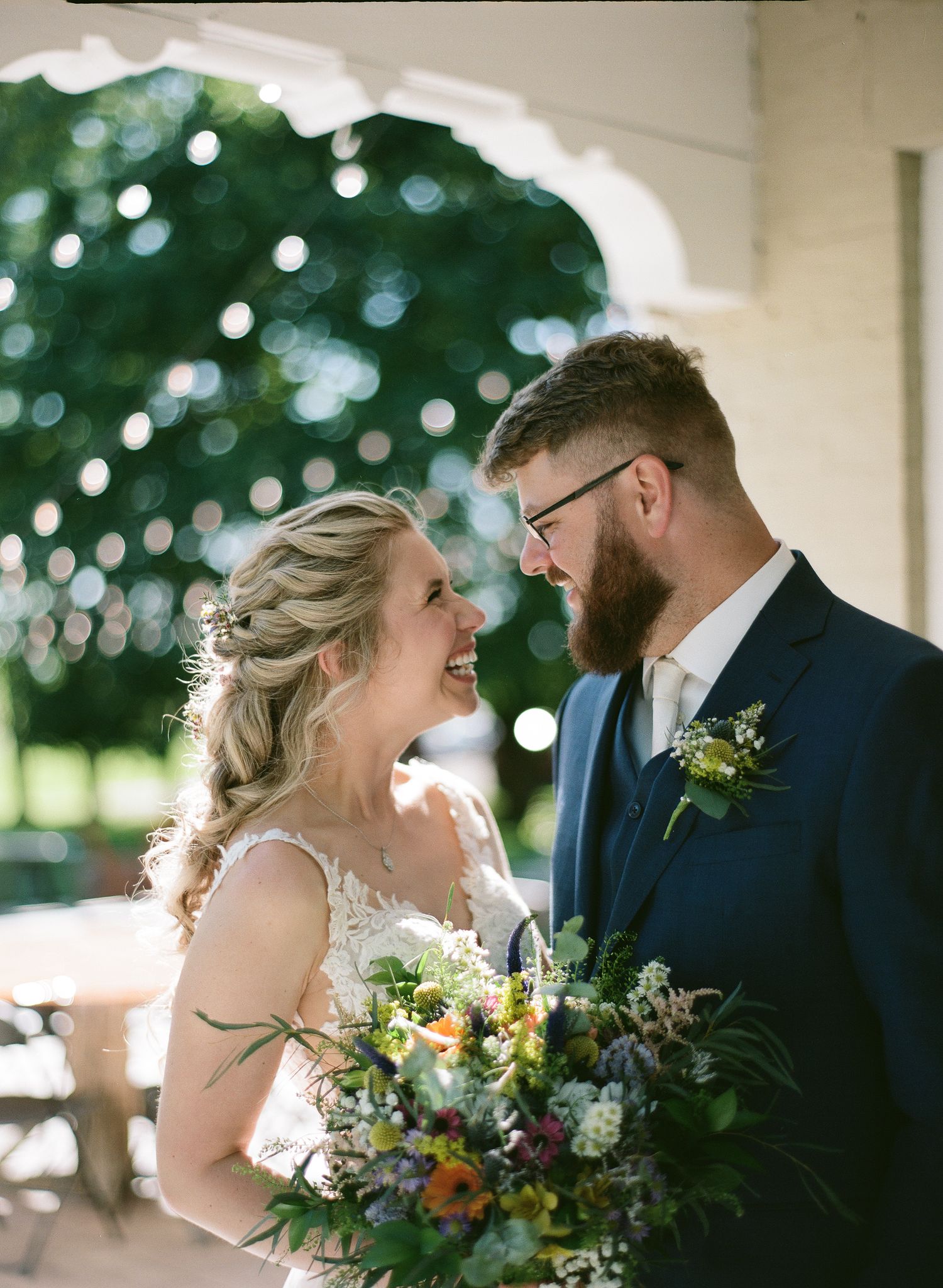 A smiling bride and bearded groom in a navy suit gaze at each other lovingly at an outdoor wedding venue.