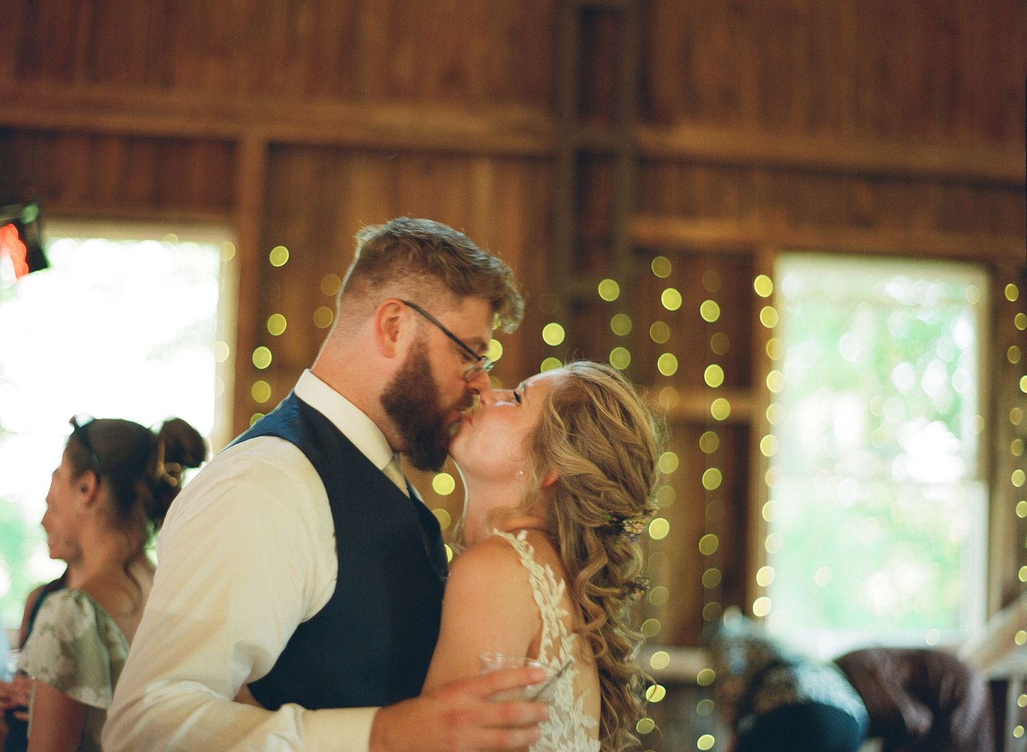 A bride and groom share a romantic first dance kiss in a warmly lit barn decorated with string lights.