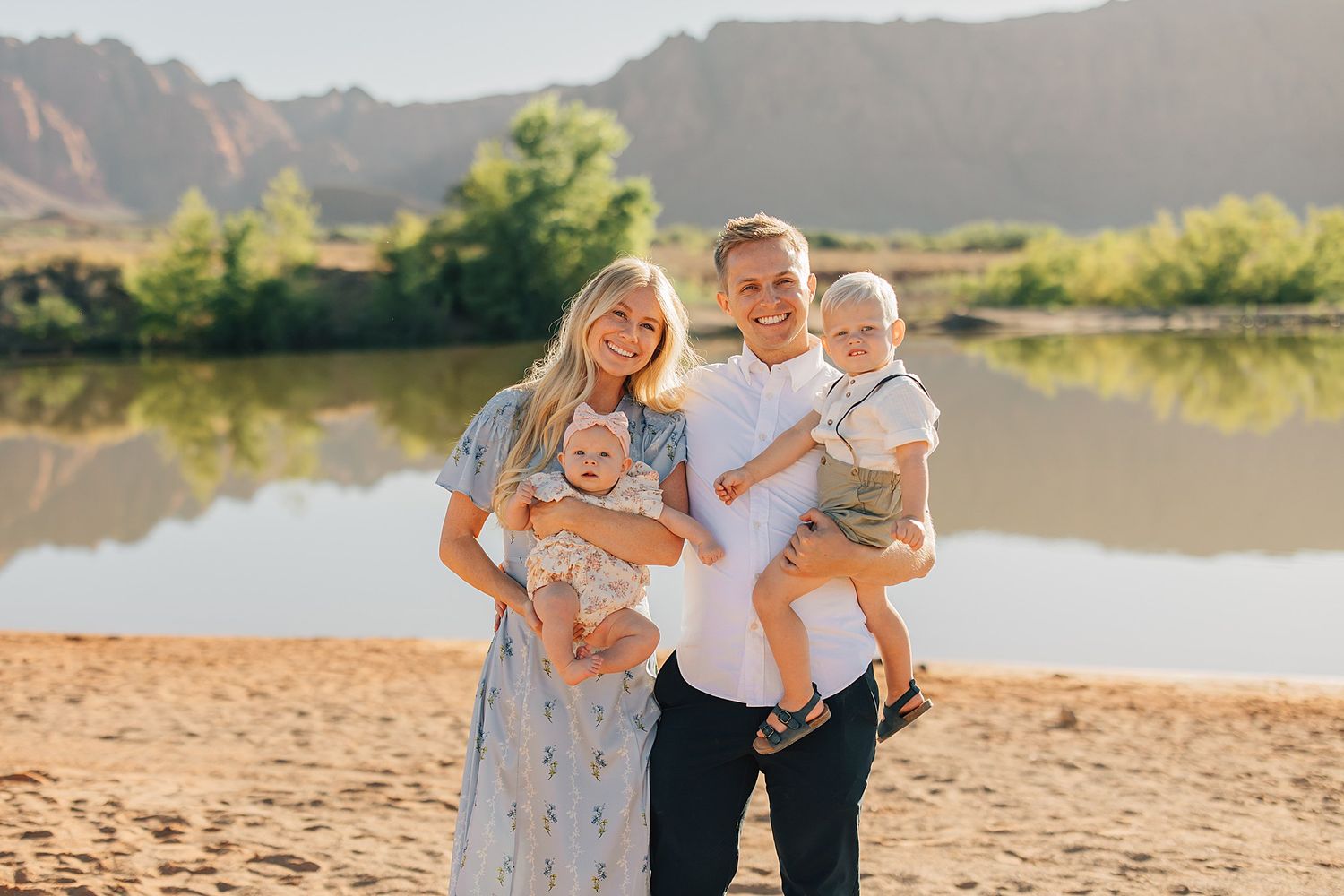 Happy family of four poses on sandy beach with scenic river and mountain backdrop in warm golden light.