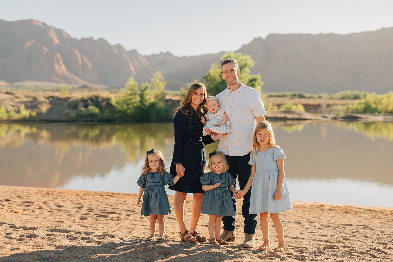 Family of six posing on sandy beach with mountain lake backdrop in warm golden light.