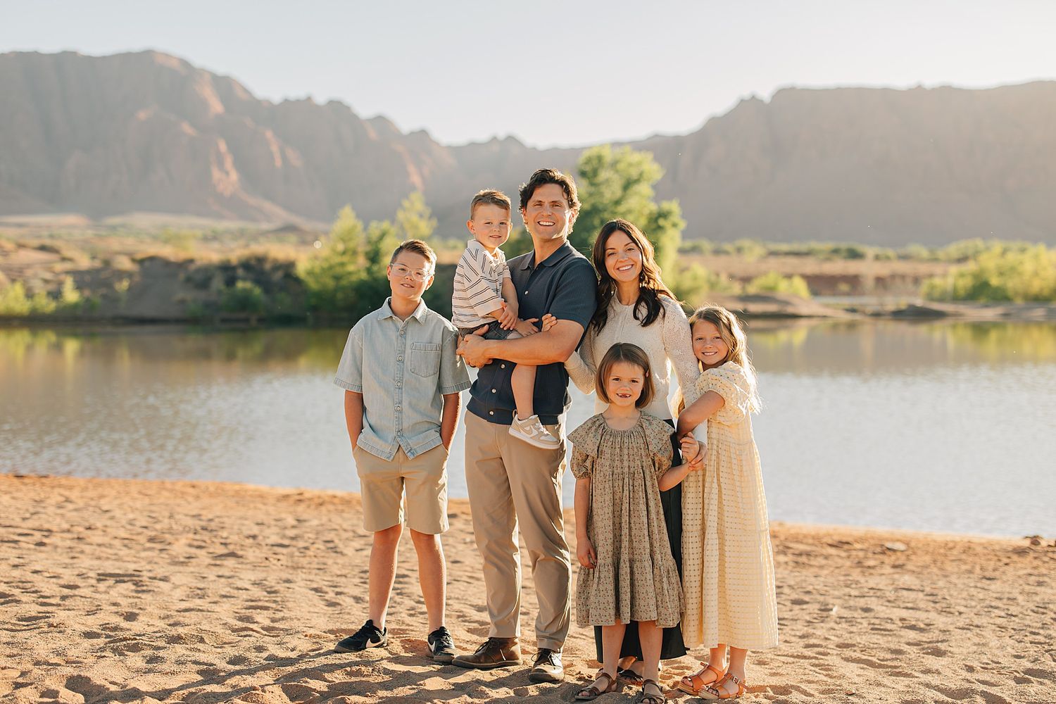 Family of six posing on sandy beach with river and red rock mountains in golden hour sunlight.