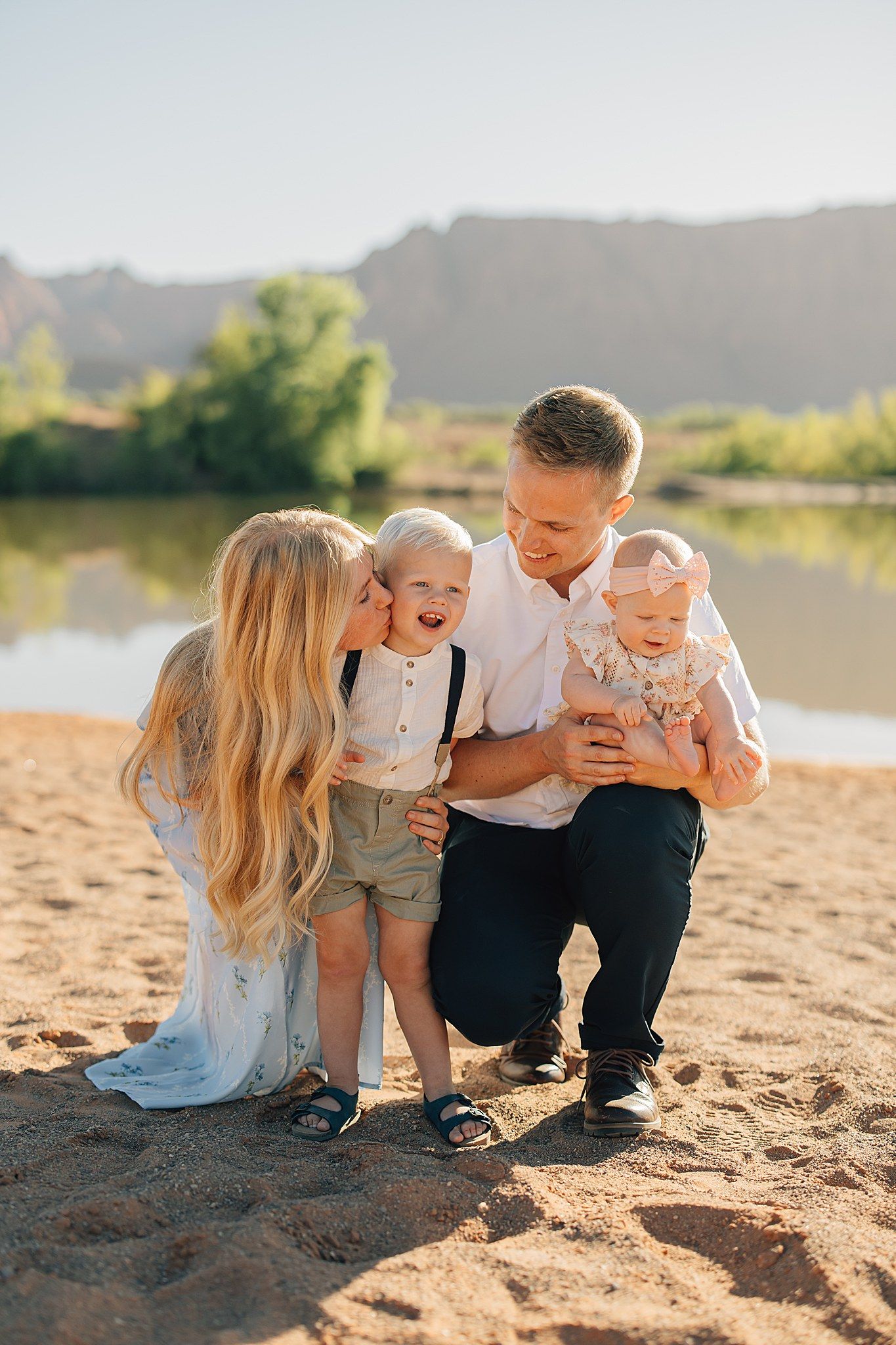 Family of four posing on sandy beach near river, mom kissing toddler son while dad holds baby girl outdoors.