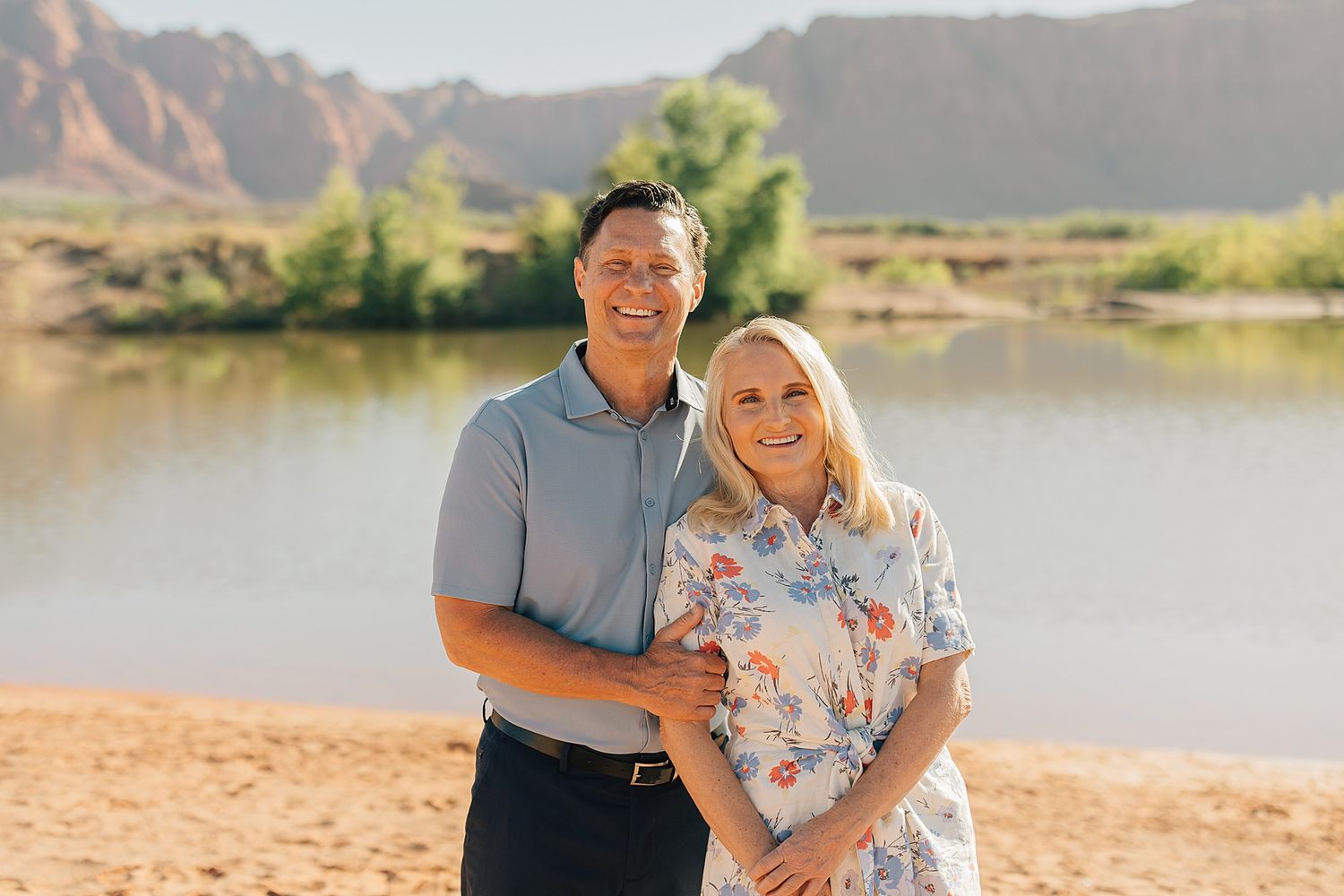 Smiling couple posing together on a sandy riverbank with red rock mountains and green trees in the background.