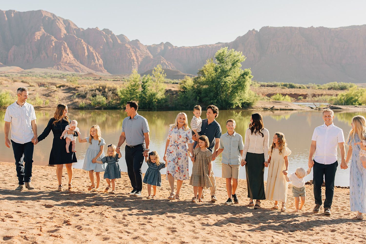 Large extended family group photo on sandy beach with red rock canyon and river backdrop on sunny day.