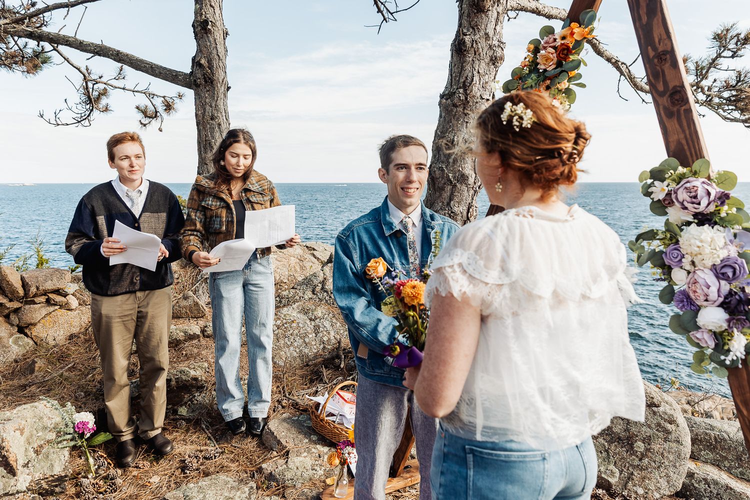 Outdoor coastal wedding ceremony with couple exchanging vows by the sea, surrounded by floral arch and pine trees.