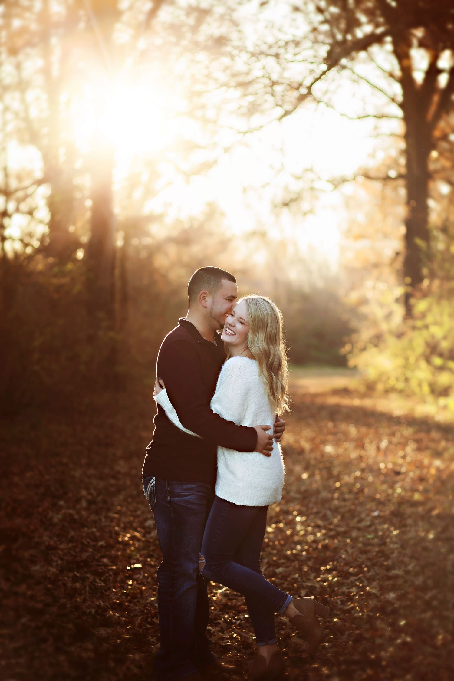 Smiling couple embracing outdoors in autumn park with golden sunlight filtering through trees, woman in white sweater, man in burgundy top.
