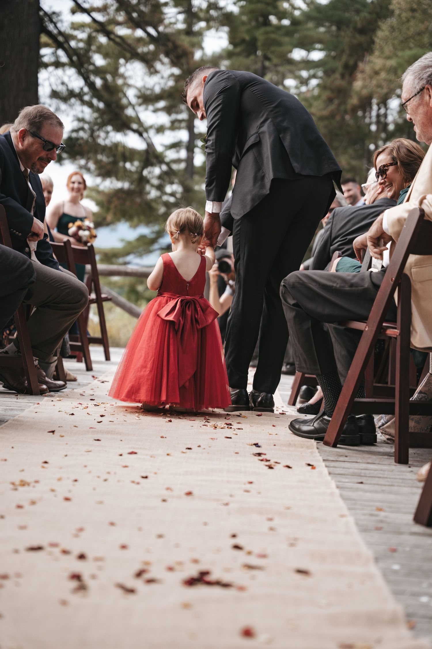 A flower girl in a red dress walks down the wedding aisle as a groomsman bends down to assist her.