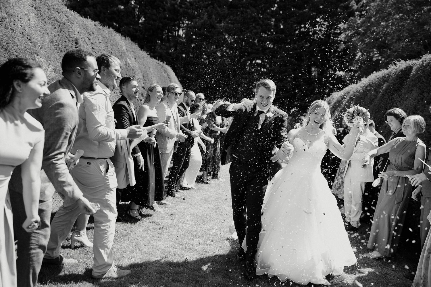Black and white wedding photo of bride and groom walking through confetti tunnel of cheering guests outdoors.