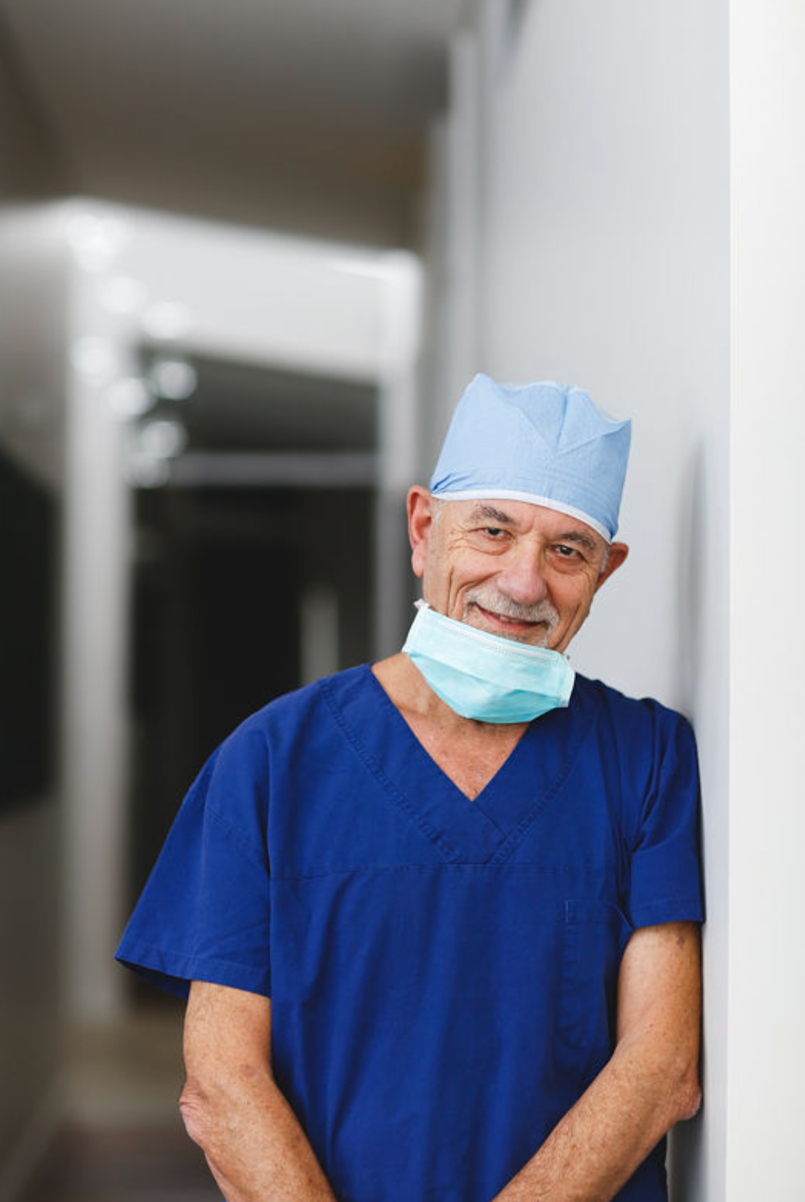 Smiling senior male surgeon in blue scrubs and surgical cap with face mask pulled down, leaning against a hospital wall.