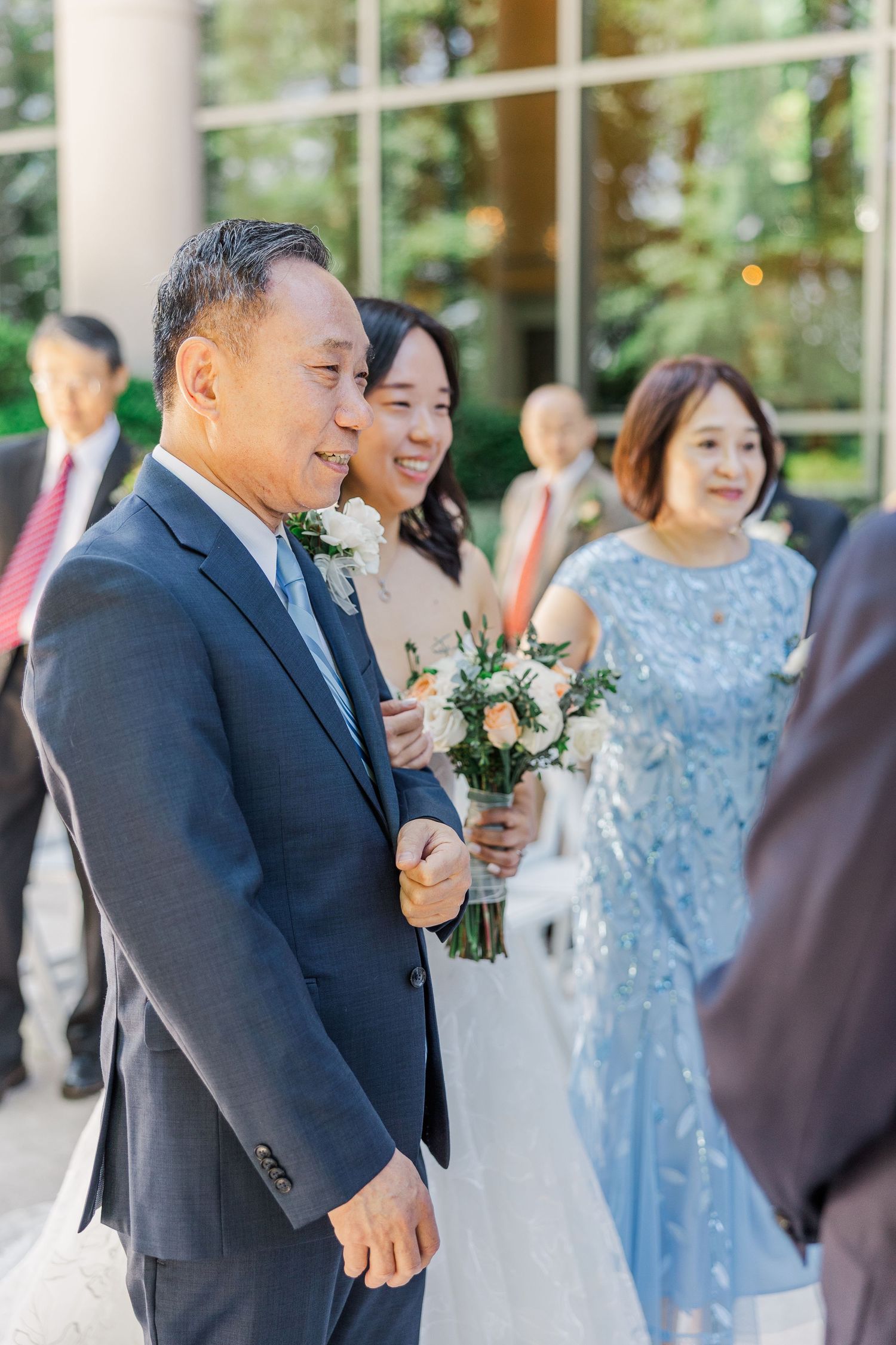 Father escorts bride down the aisle at outdoor wedding ceremony, with guests and mother in blue dress following behind.