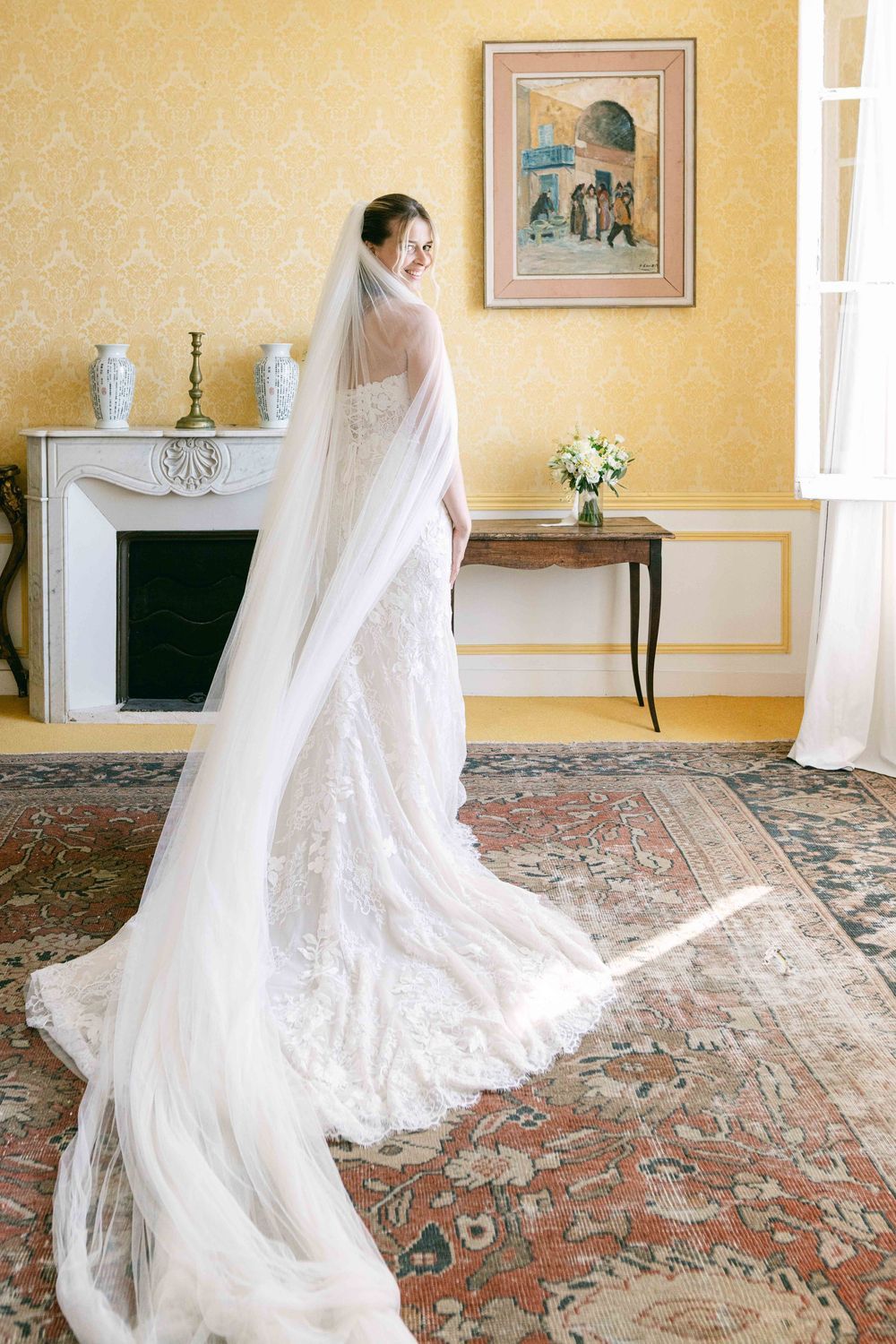 Bride in elegant lace wedding gown with long cathedral veil stands in a yellow room with fireplace and floral bouquet.