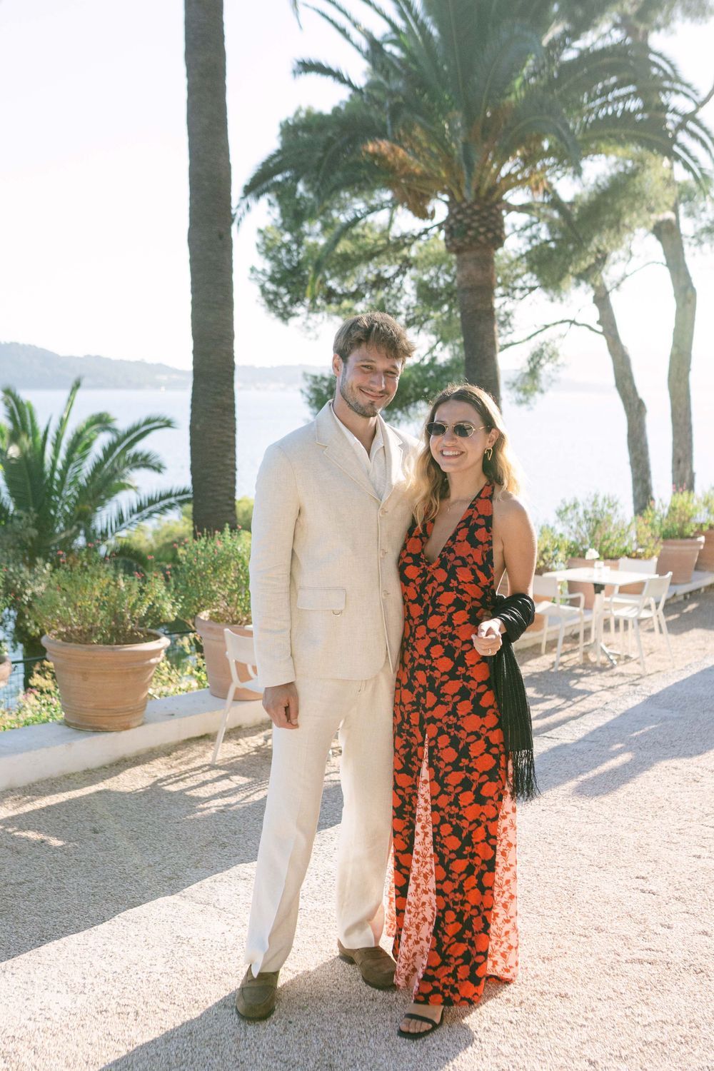 Couple posing outdoors in elegant attire near palm trees and ocean, woman in red floral dress, man in cream suit.