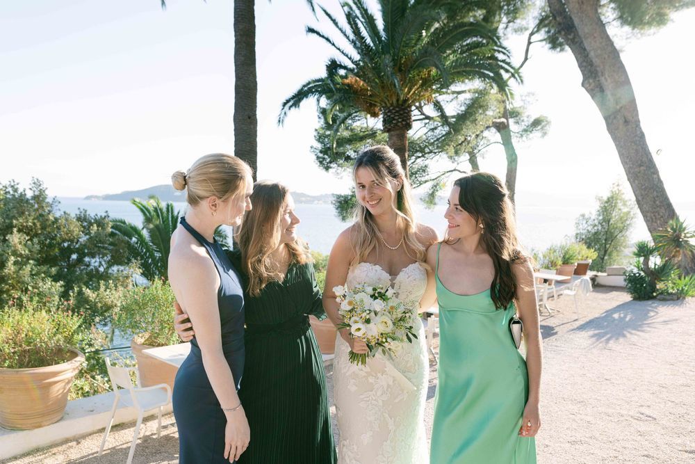 Bride in strapless lace gown holding bouquet laughing with bridesmaids in green and navy dresses outdoors.