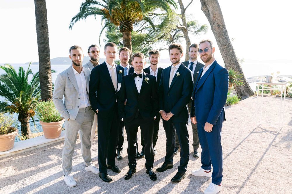 Groom in black tuxedo poses with groomsmen in suits outdoors near palm trees at a sunny wedding venue.